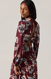 A woman is wearing a long-sleeved dress with a floral pattern in shades of blue, beige, and white on a maroon background. The dress features a cutout on the back with a tie detail at the waist, creating a stylish and elegant look.